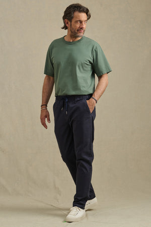 A man stands slightly turned, wearing a green tee, Velvet by Graham & Spencer's JESSE PANT in dark blue, and white sneakers against a plain beige background.