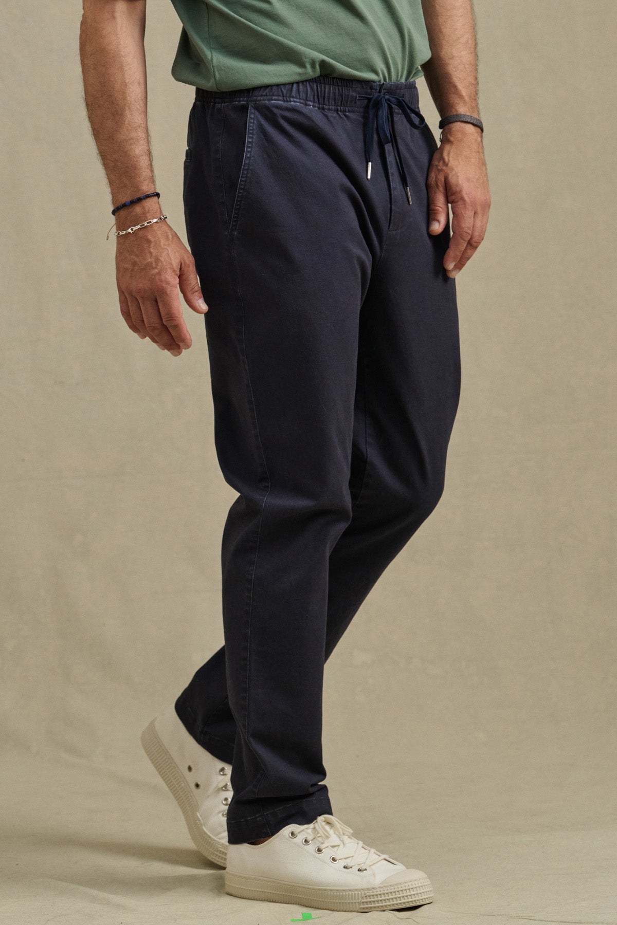 A man wearing a green shirt, white sneakers, and Velvet by Graham & Spencer’s JESSE PANT in navy stands on a beige background.-41009007657153