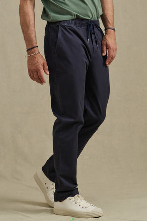 A man wearing a green shirt, white sneakers, and Velvet by Graham & Spencer’s JESSE PANT in navy stands on a beige background.