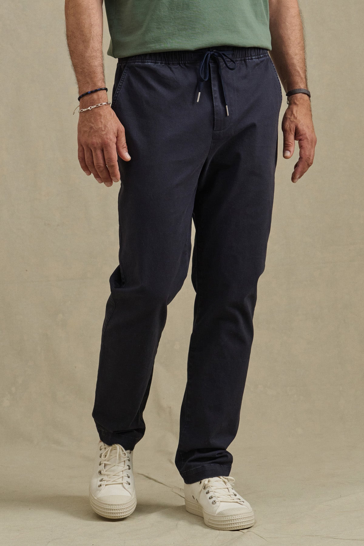 A person wearing a green T-shirt, Velvet by Graham & Spencer’s navy JESSE PANT, and white sneakers stands against a plain beige background.-41009007624385