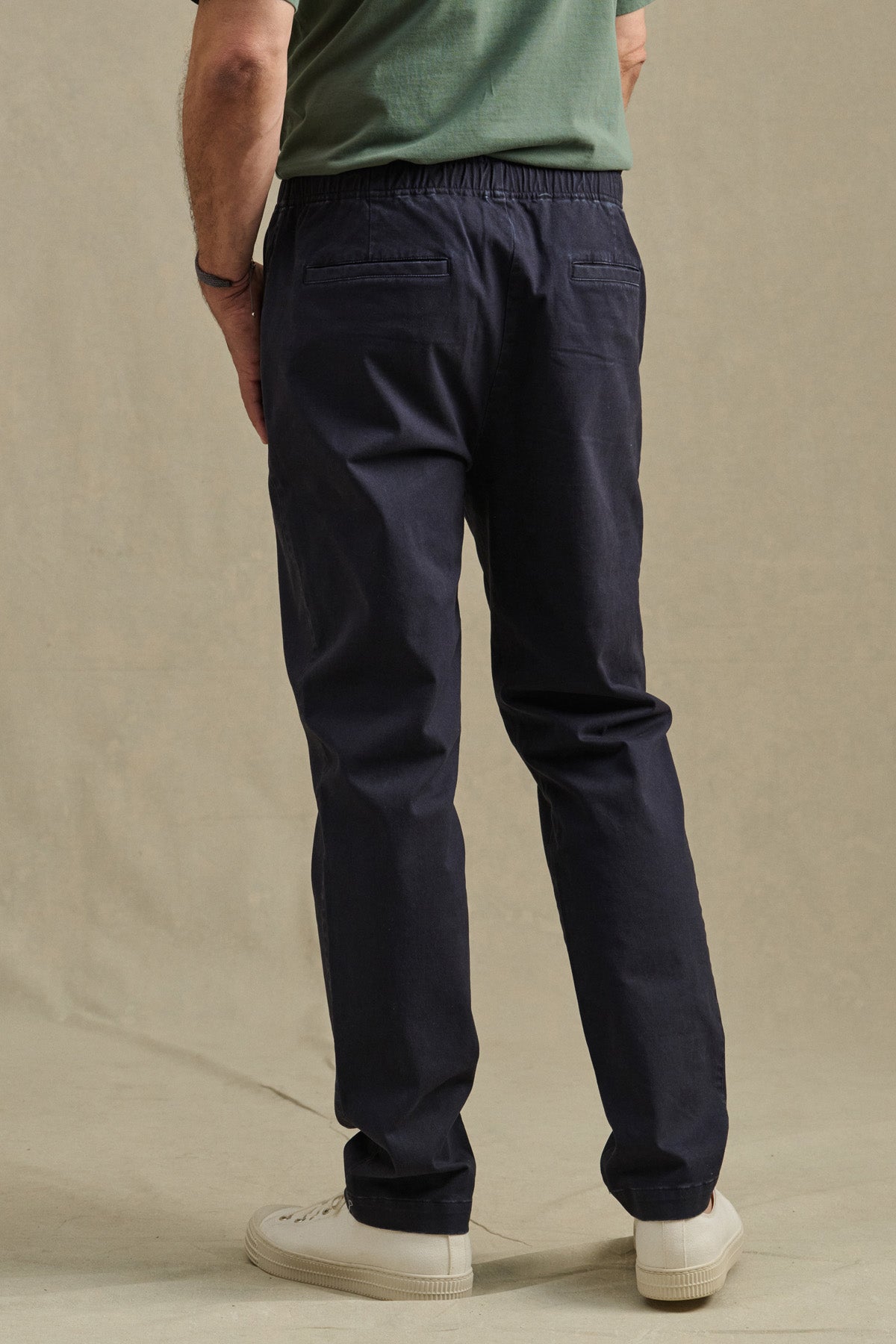 Wearing a green shirt, Velvet by Graham & Spencer's JESSE PANT—tailored fit cotton stretch pants with a drawstring waist—and white sneakers, a person stands on a beige background facing away from the camera.