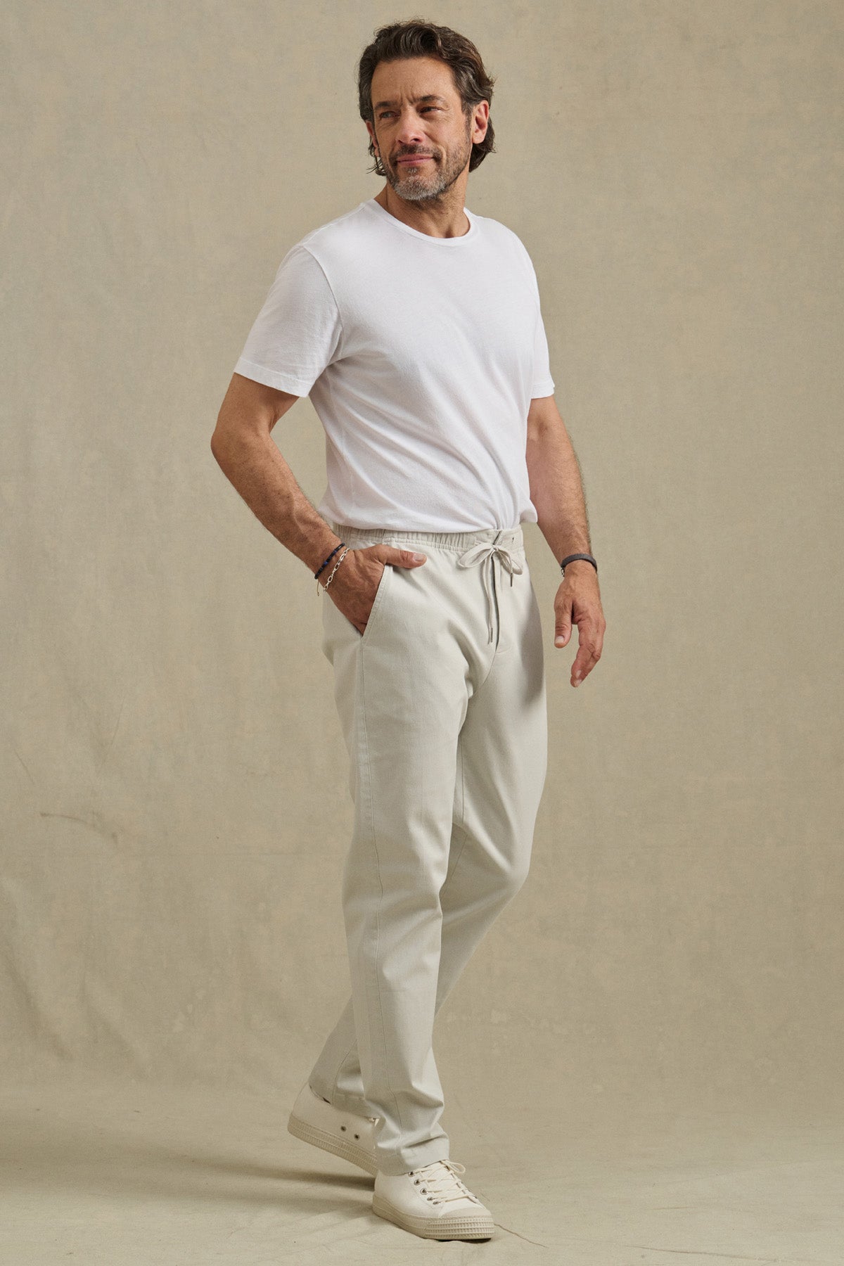 A man stands before a beige backdrop wearing a white t-shirt, white sneakers, and the JESSE PANT by Velvet by Graham & Spencer—modern fit cotton-stretch pants with a drawstring elastic waist, one hand in his pocket.-41338469580993