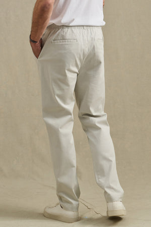 A person is seen from behind with one hand in their pocket, wearing the JESSE PANT by Velvet by Graham & Spencer in light beige cotton stretch, styled with a white shirt and white sneakers.