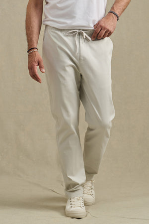 A person stands on a neutral background wearing the Velvet by Graham & Spencer JESSE PANT in light beige cotton stretch with a drawstring waist, paired with a white T-shirt and white sneakers.