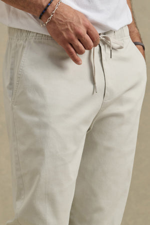 A person wearing the JESSE PANT by Velvet by Graham & Spencer—modern fit, light beige cotton stretch pants with a drawstring elastic waist—and a white shirt, one hand resting near the waistband.