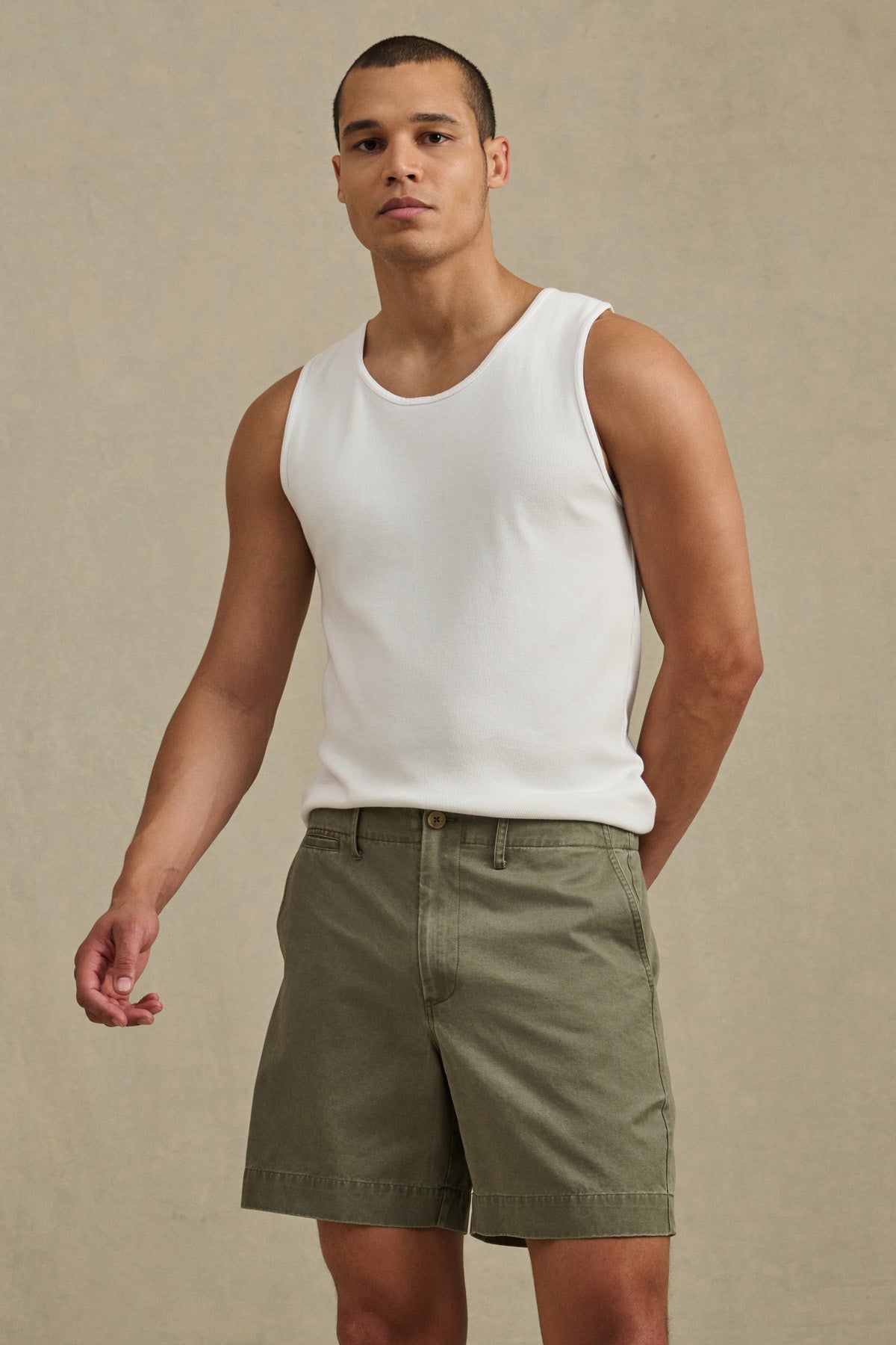 A man stands against a neutral background wearing a white sleeveless tank and Velvet by Graham & Spencer JOEL SHORT, olive green 5-pocket shorts, with his left hand behind his back.-41338470006977