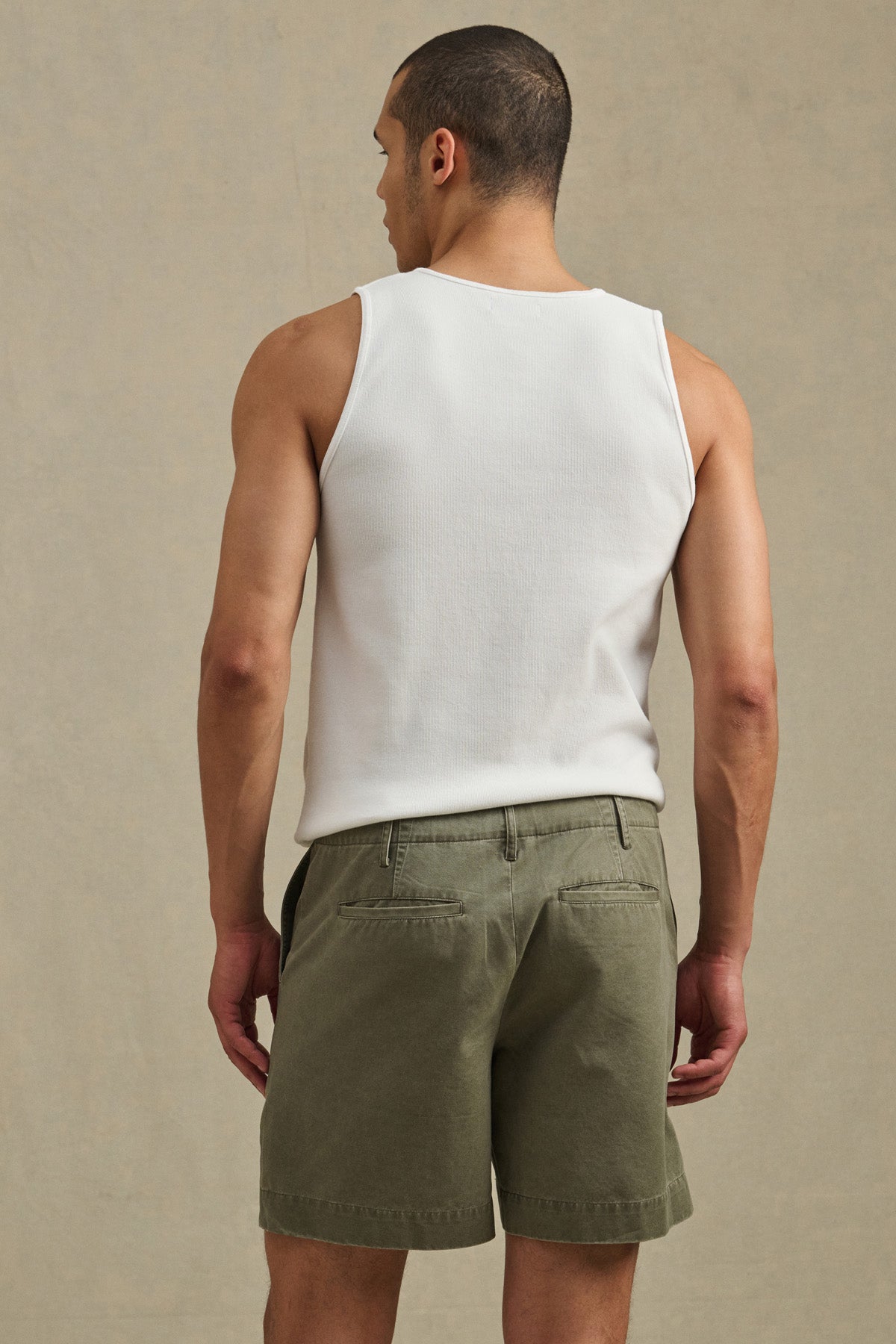 A man stands facing away, wearing a white tank top and Olive JOEL SHORT 5-pocket shorts by Velvet by Graham & Spencer against a plain, neutral background.