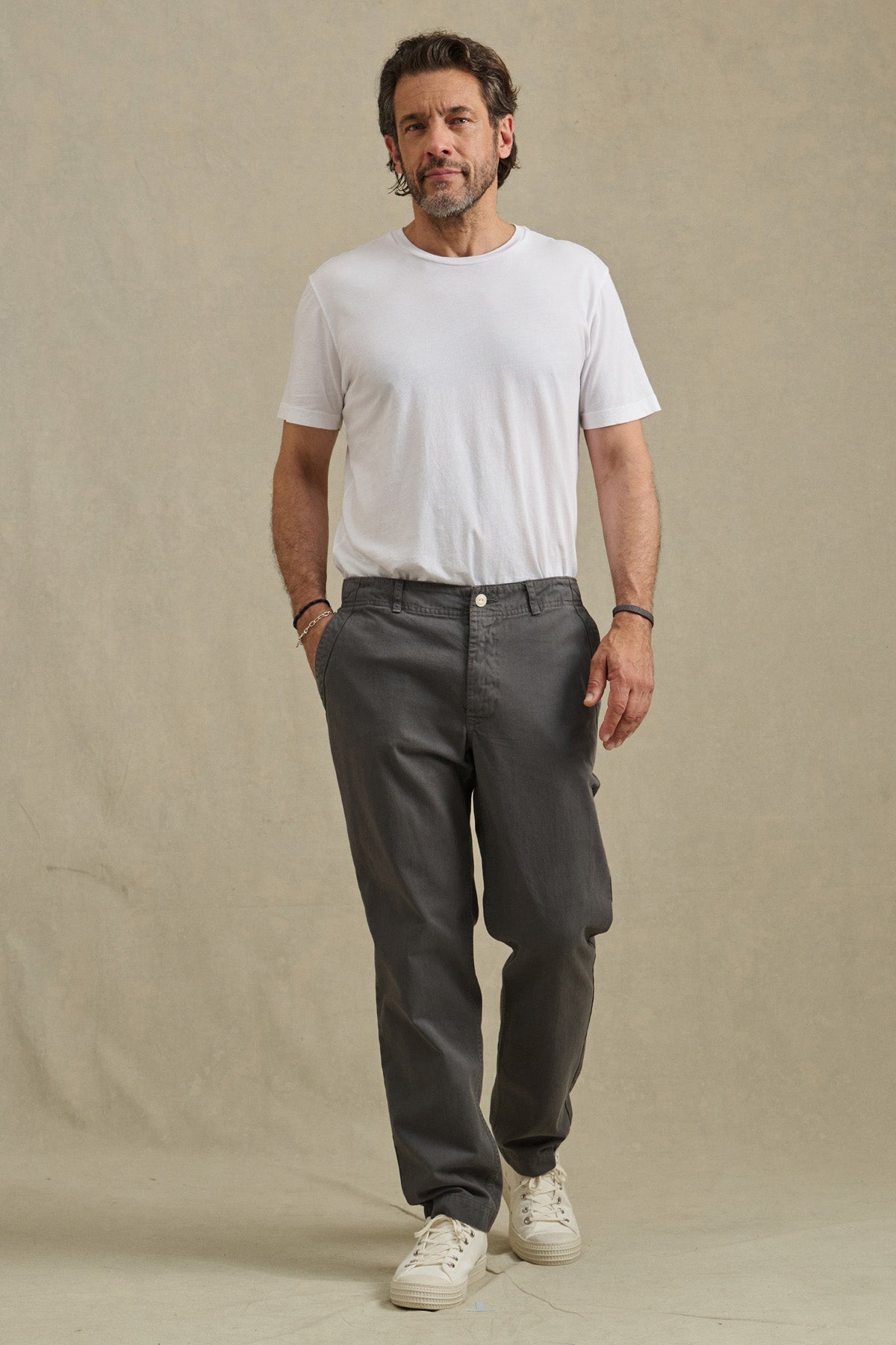 A man stands against a plain background wearing a white t-shirt and the LENNOX PANT by Velvet by Graham & Spencer in relaxed fit gray cotton twill, paired with white sneakers—an ideal look for everyday wear.