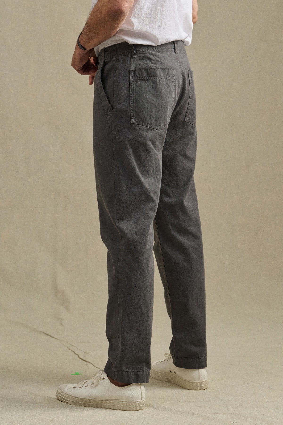 A person in a white T-shirt, Velvet by Graham & Spencer LENNOX PANT in relaxed fit gray cotton twill, and white sneakers stands on a beige surface facing away from the camera—an easy everyday look.