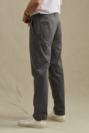 A person in a white T-shirt, Velvet by Graham & Spencer LENNOX PANT in relaxed fit gray cotton twill, and white sneakers stands on a beige surface facing away from the camera—an easy everyday look.