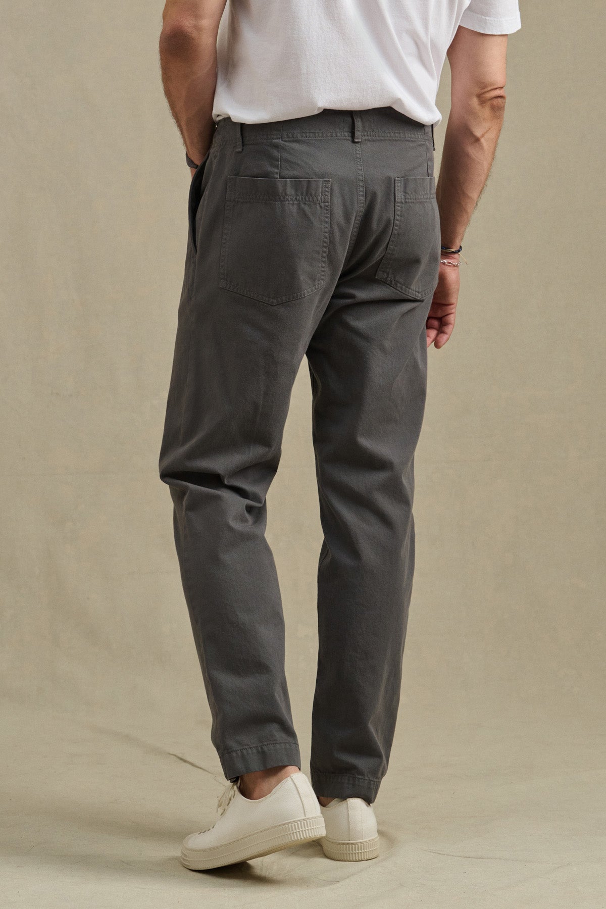 A man stands with his back to the camera in a white t-shirt, Velvet by Graham & Spencer LENNOX PANT (relaxed fit, gray cotton twill), and white sneakers against a beige background—ideal for everyday wear.