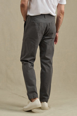 A man stands with his back to the camera in a white t-shirt, Velvet by Graham & Spencer LENNOX PANT (relaxed fit, gray cotton twill), and white sneakers against a beige background—ideal for everyday wear.