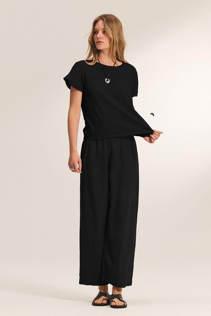 A woman stands against a plain background wearing a black short-sleeve top, Velvet by Graham & Spencer's LOLA LINEN PANT with an elastic waist, black sandals, and a pendant necklace.