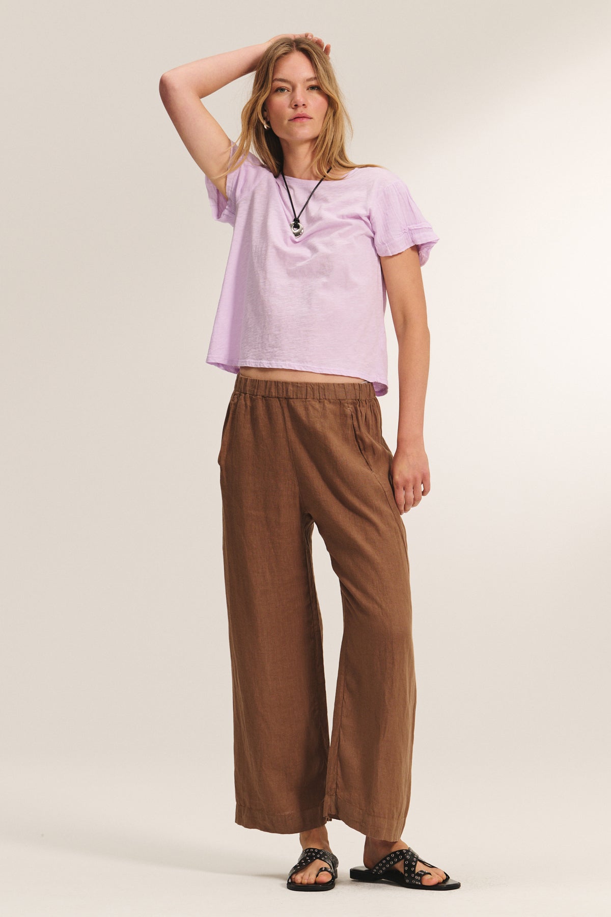   A woman poses against a plain background wearing the PATRICIA TEE by Velvet by Graham & Spencer in light purple, paired with brown wide-leg pants, black sandals, and a necklace, one hand resting on her head. 