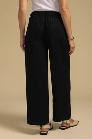 A person viewed from the back wearing the PENELOPE LINEN PANT, designed with cropped, wide-leg linen trousers and an elasticized waistband.