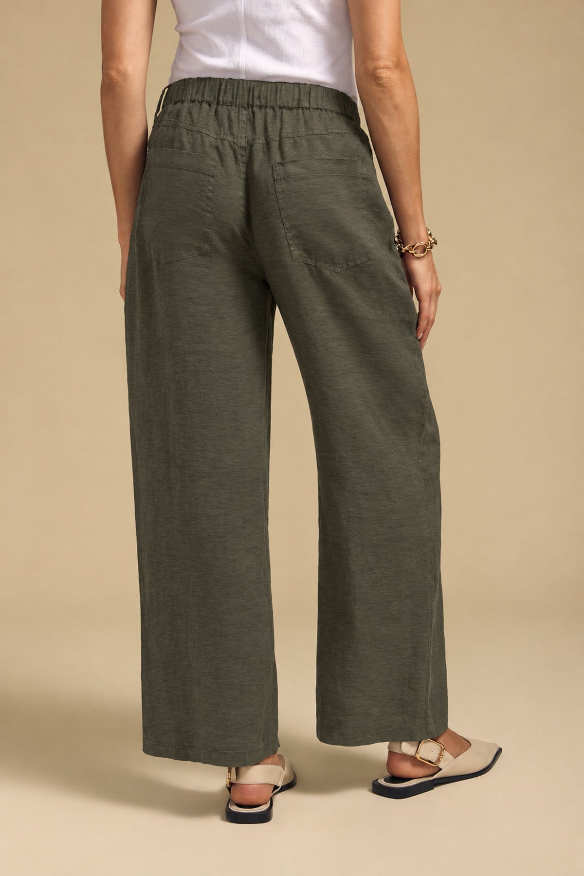 Rear view of a person wearing olive green PENELOPE LINEN PANT wide-leg linen pants with an elastic waistband and flat slingback sandals.