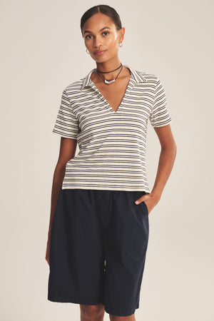 A woman in the PERRIN POLO by Velvet by Graham & Spencer, featuring yarn-dyed stripes and short sleeves, pairs it with navy knee-length shorts and stands with one hand in her pocket against a plain background.