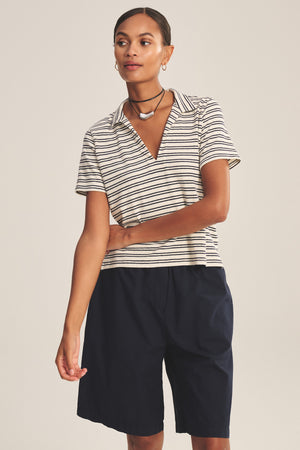 A woman wears the Velvet by Graham & Spencer PERRIN POLO, a yarn-dyed stripe cropped polo, paired with navy knee-length shorts against a plain background.