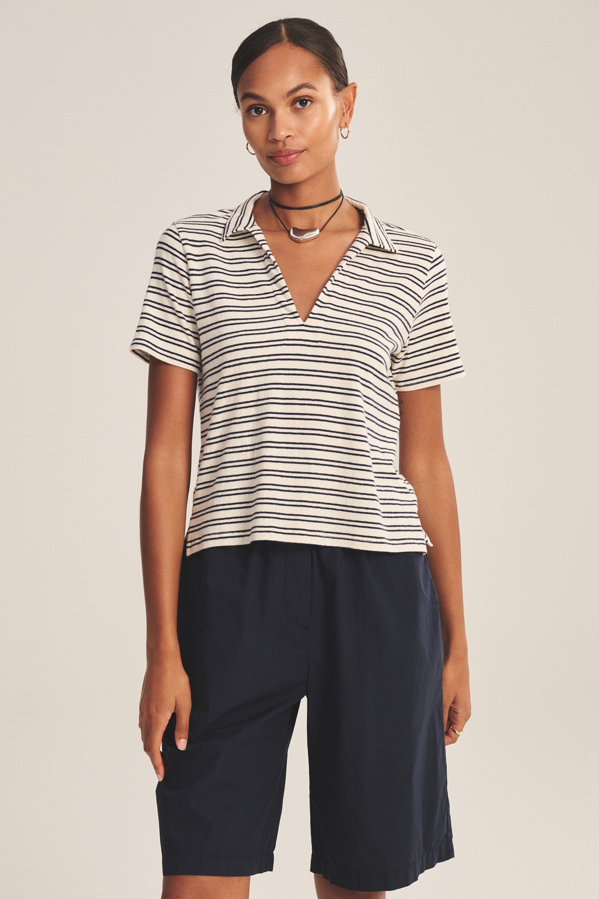 A woman faces forward wearing the Velvet by Graham & Spencer PERRIN POLO, styled with knee-length black shorts, a choker necklace, and hoop earrings.-41089063059649