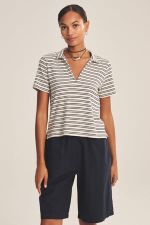 A woman faces forward wearing the Velvet by Graham & Spencer PERRIN POLO, styled with knee-length black shorts, a choker necklace, and hoop earrings.