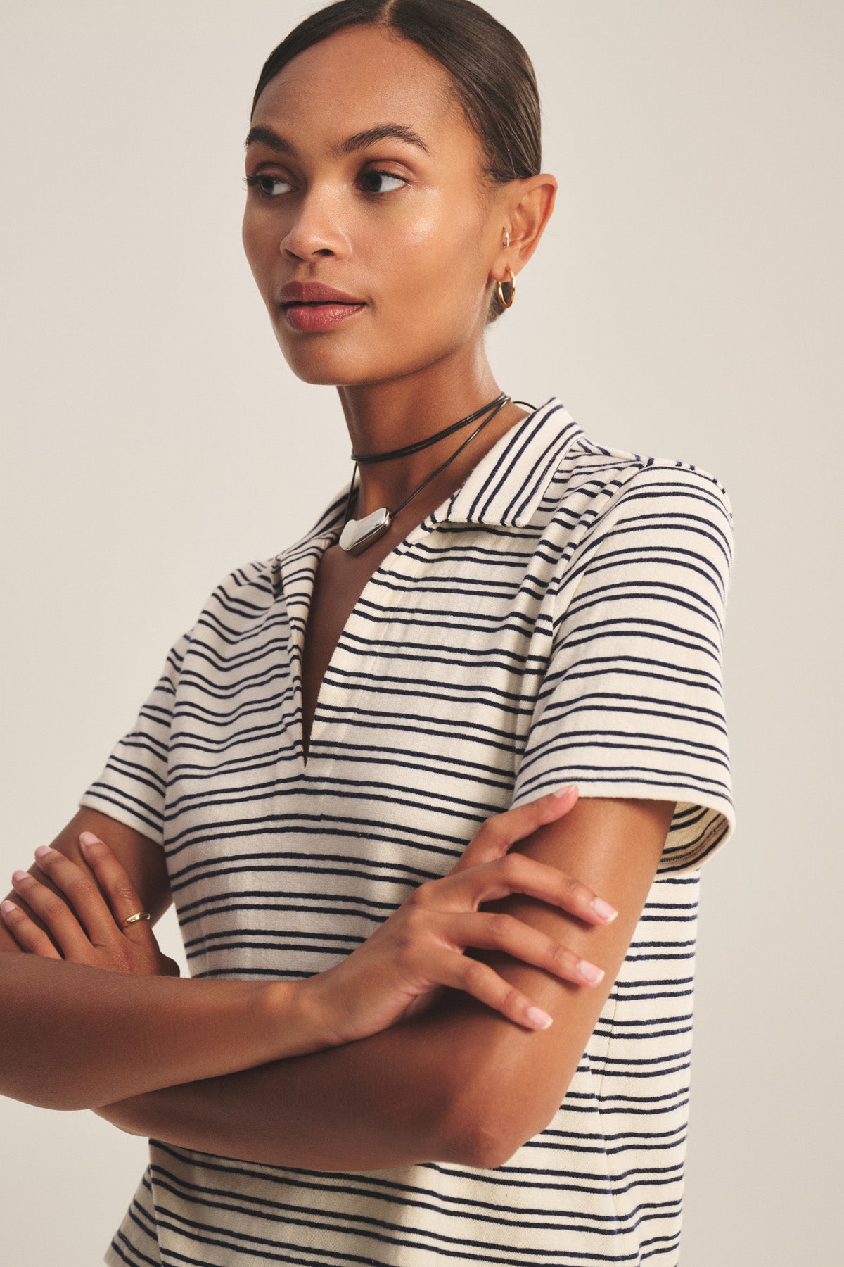 A woman stands with arms crossed, looking to the side, wearing the Velvet by Graham & Spencer PERRIN POLO—a cropped, yarn-dyed stripe polo with short sleeves—paired with a pendant necklace and hoop earrings.-41089063026881