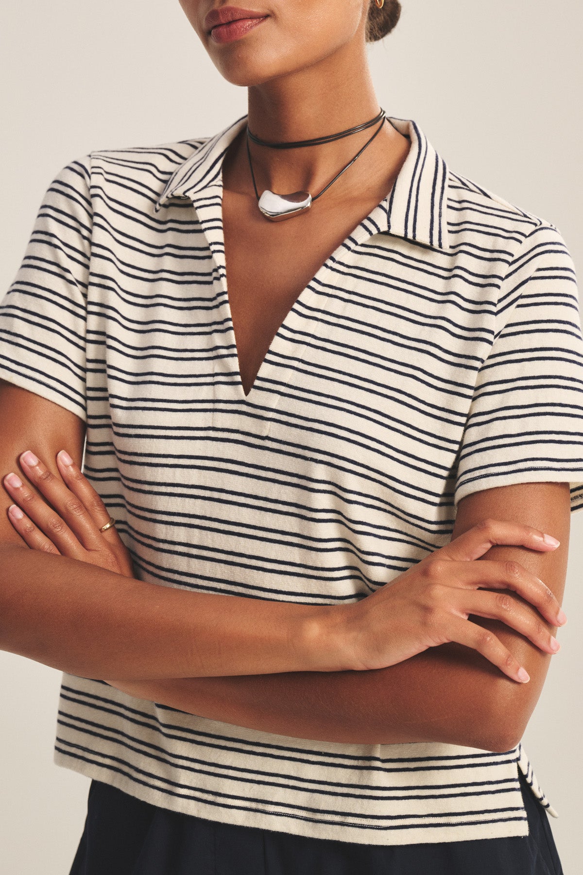 A woman with arms crossed wears the PERRIN POLO by Velvet by Graham & Spencer, a yarn-dyed stripe polo with short sleeves, collared V-neckline, and a layered black necklace featuring a white pendant.
