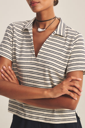 A woman with arms crossed wears the PERRIN POLO by Velvet by Graham & Spencer, a yarn-dyed stripe polo with short sleeves, collared V-neckline, and a layered black necklace featuring a white pendant.