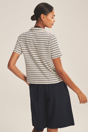 A person stands with their back turned, wearing the Velvet by Graham & Spencer PERRIN POLO in black and white stripes and navy shorts against a neutral background.