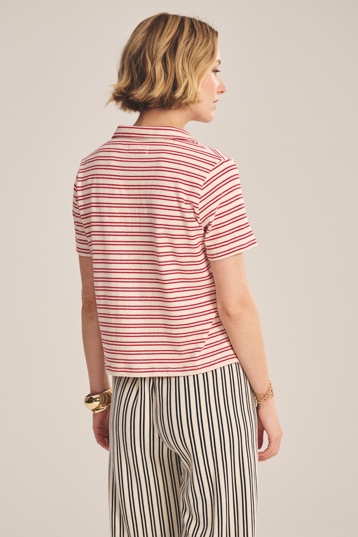 A person stands facing away, wearing the Velvet by Graham & Spencer PERRIN POLO, a red and white V-neck short-sleeve T-shirt, paired with black and white vertically striped pants.