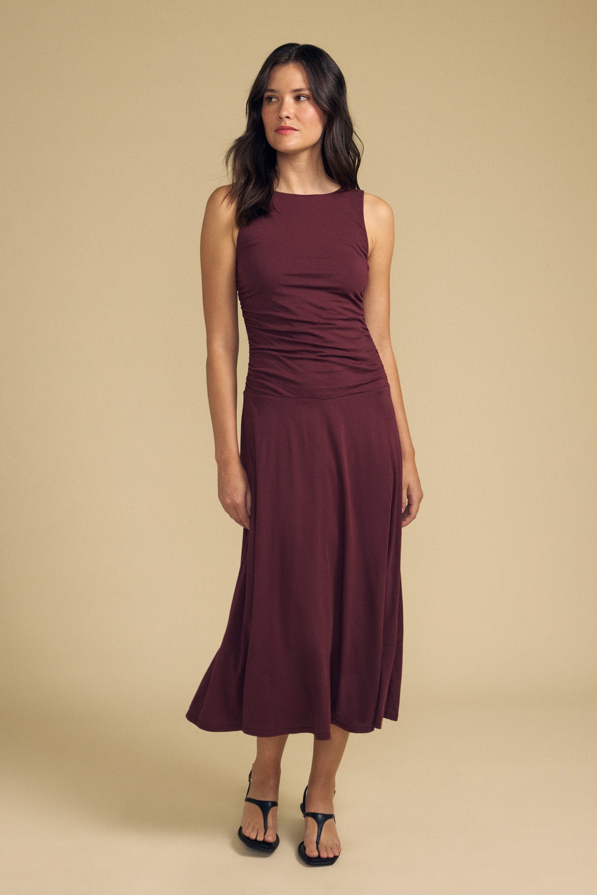 A woman stands against a beige background, wearing the Velvet by Graham & Spencer PIA DRESS—a sleeveless maroon midi dress with a delicate, gauzy whisper—and black sandals, gazing slightly to the side.-41838201569473