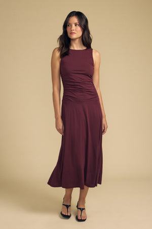 A woman stands against a beige background, wearing the Velvet by Graham & Spencer PIA DRESS—a sleeveless maroon midi dress with a delicate, gauzy whisper—and black sandals, gazing slightly to the side.