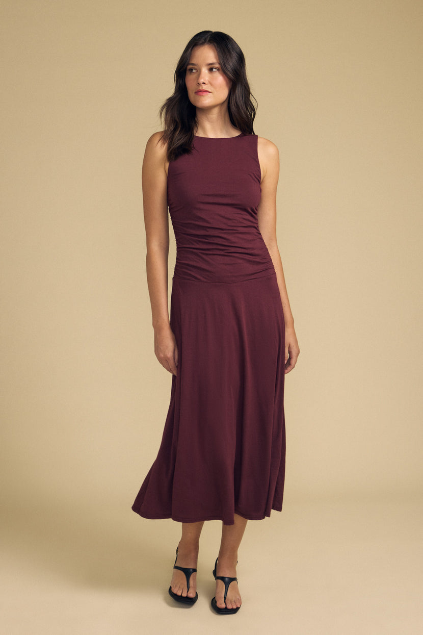 A woman stands against a beige background, wearing the Velvet by Graham & Spencer PIA DRESS—a sleeveless maroon midi dress with a delicate, gauzy whisper—and black sandals, gazing slightly to the side.