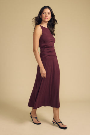 A woman wearing the PIA DRESS by Velvet by Graham & Spencer—a sleeveless, burgundy-lined midi dress—pairs it with black heeled sandals as she stands against a plain beige background.