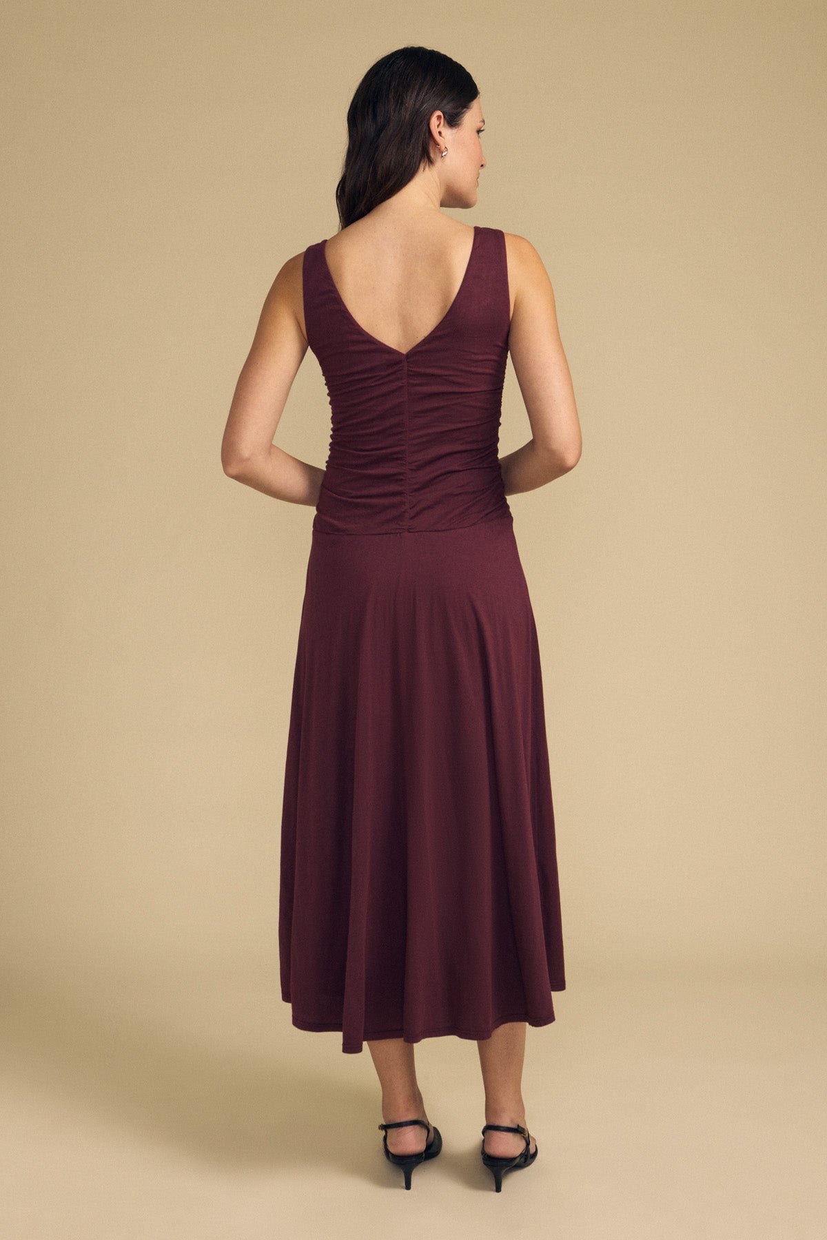 A woman stands with her back to the camera against a plain beige background, wearing the PIA DRESS—a sleeveless, burgundy lined midi dress from Velvet by Graham & Spencer—paired with black heels.