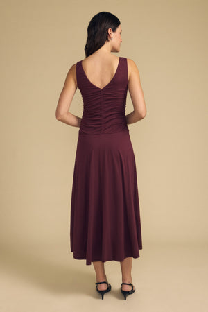 A woman stands with her back to the camera against a plain beige background, wearing the PIA DRESS—a sleeveless, burgundy lined midi dress from Velvet by Graham & Spencer—paired with black heels.