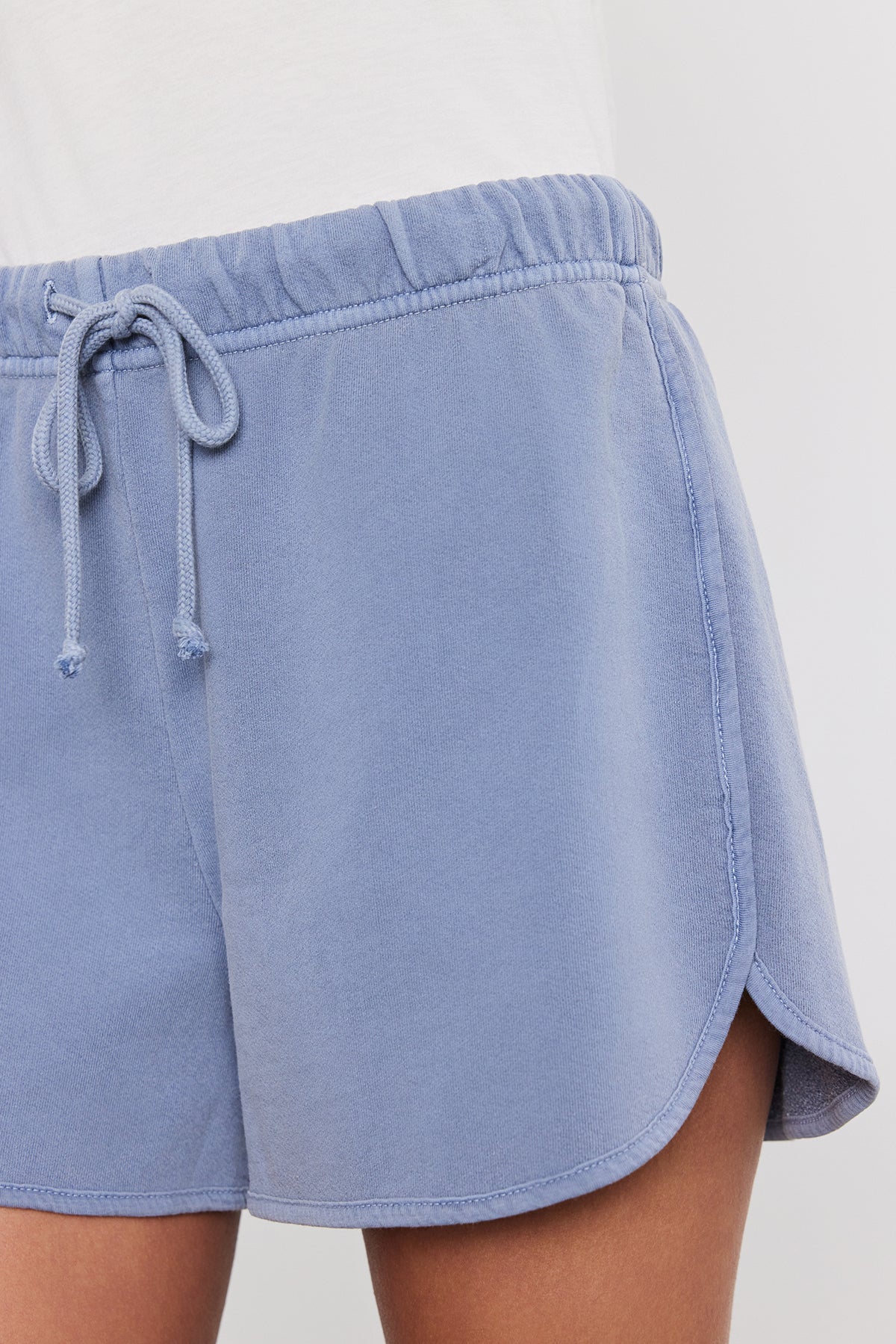   Close-up of a person wearing light blue drawstring shorts and a white top, showing the waist and upper thigh area. These relaxed fit, classic gym shorts are perfect warm weather basics. The PRESELY SHORT by Velvet by Graham & Spencer is a great addition to any summer wardrobe. 