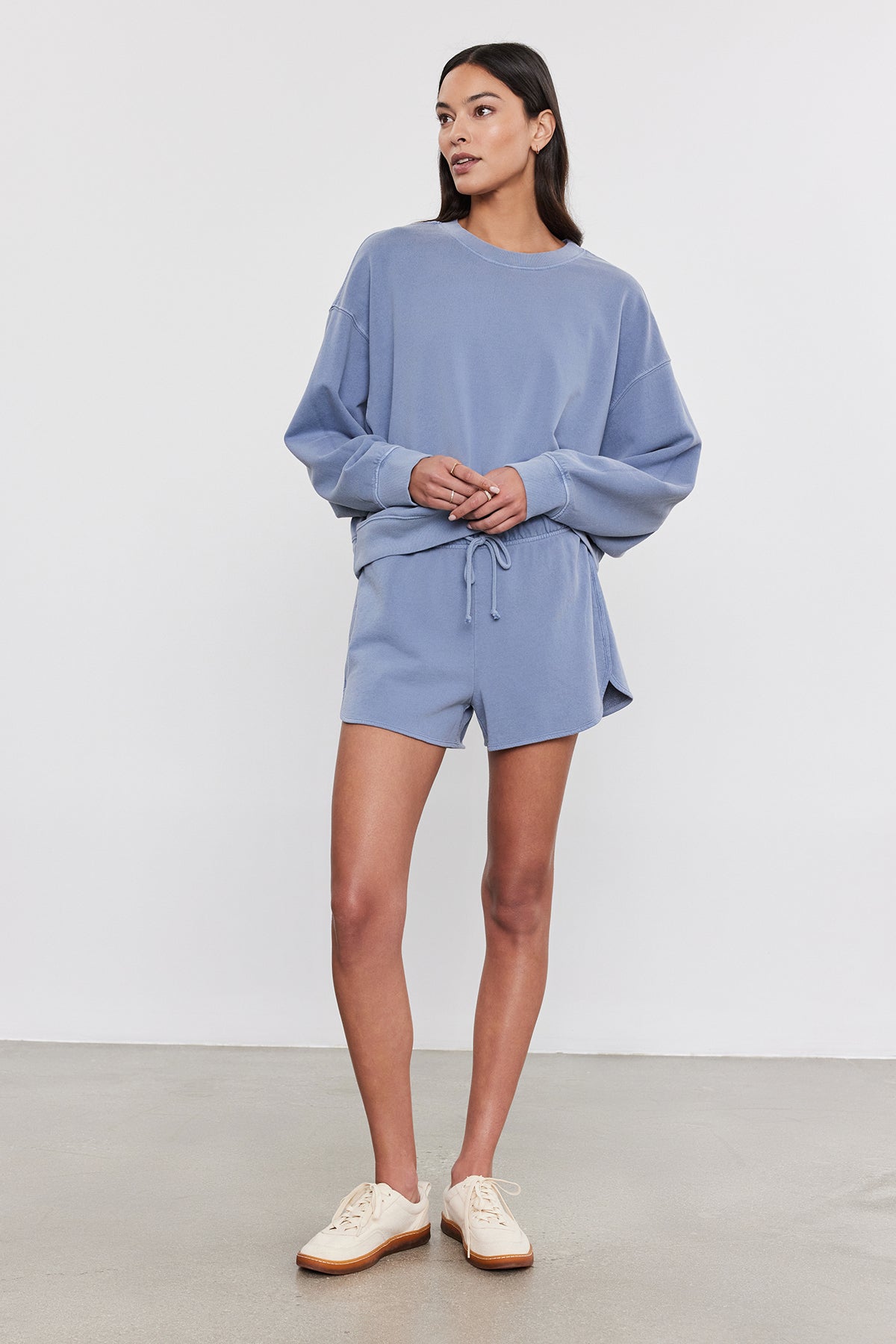   A person stands against a plain background wearing a light blue, vintage-inspired SKYLAR SWEATSHIRT and matching shorts made from soft French terry cotton by Velvet by Graham & Spencer, paired with white sneakers. 