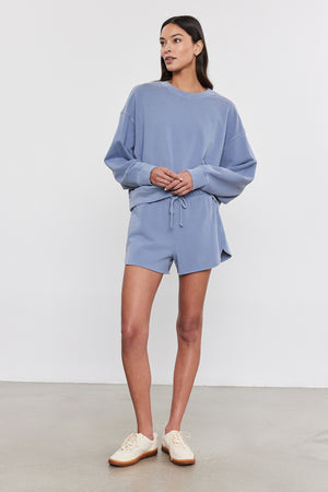 A person stands against a plain background wearing a light blue, vintage-inspired SKYLAR SWEATSHIRT and matching shorts made from soft French terry cotton by Velvet by Graham & Spencer, paired with white sneakers.