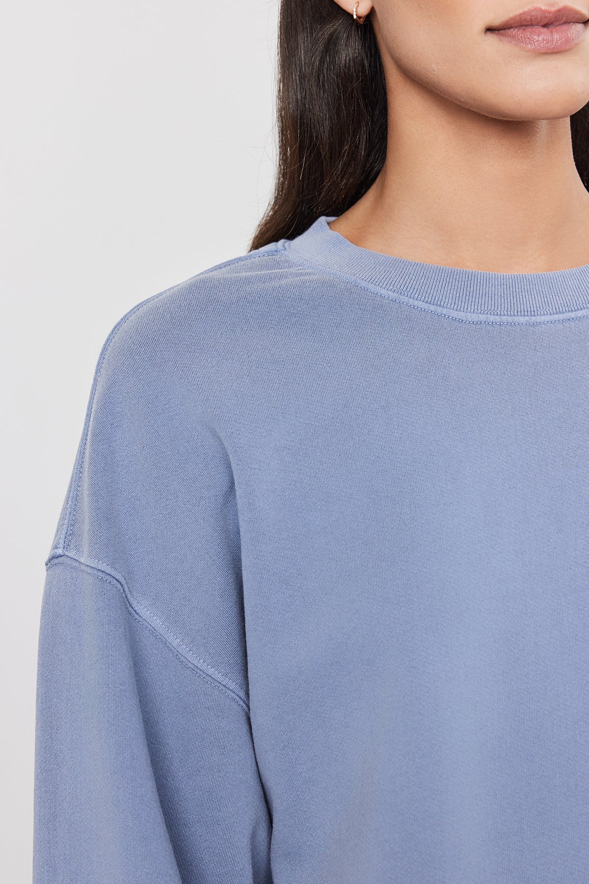   A person wearing a light blue, vintage-inspired SKYLAR SWEATSHIRT by Velvet by Graham & Spencer. The relaxed fit highlights the lower half of their face and upper torso, crafted from soft French terry cotton. 