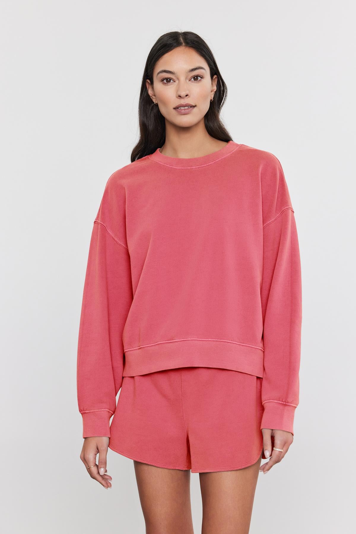 A woman wearing a coral pink, pigment-dyed Velvet by Graham & Spencer SKYLAR SWEATSHIRT and matching shorts standing against a white background.-36910094516417