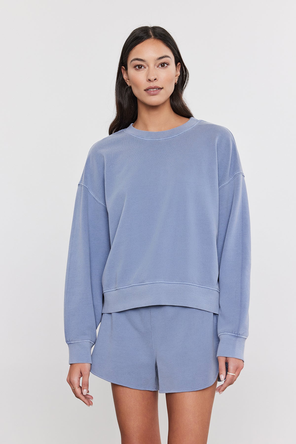   A person with long hair is wearing a relaxed fit, light blue SKYLAR SWEATSHIRT by Velvet by Graham & Spencer and matching shorts made from vintage-inspired French terry cotton. They are standing against a plain, light grey background. 