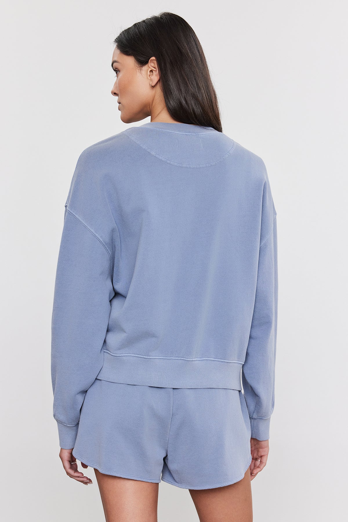   A person with long dark hair is facing away from the camera, wearing a light blue SKYLAR SWEATSHIRT by Velvet by Graham & Spencer and matching shorts in a relaxed fit against a white background. 