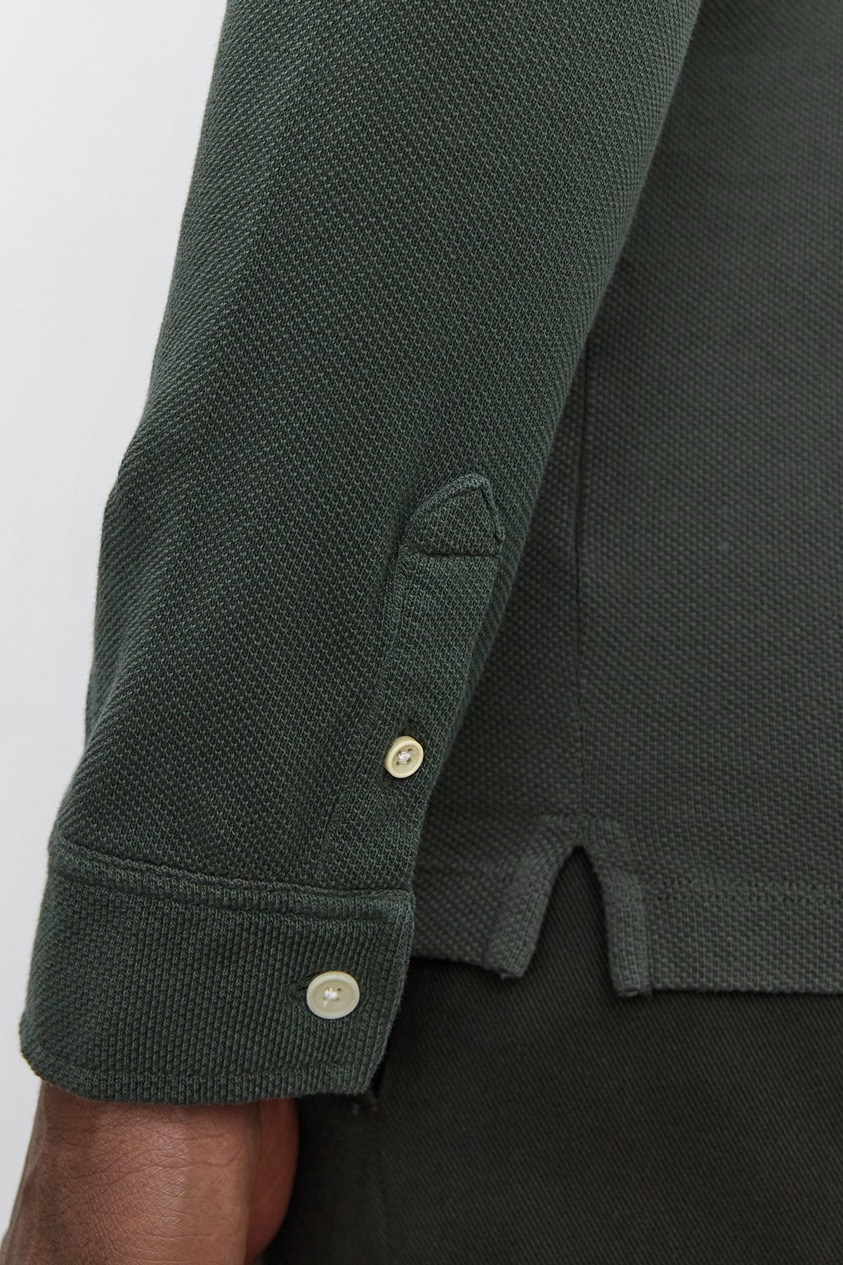   Close-up of a person wearing the MIKE POLO by Velvet by Graham & Spencer, a dark green piqué cotton long-sleeve shirt featuring textured fabric and buttoned cuffs, with visible side slits. 