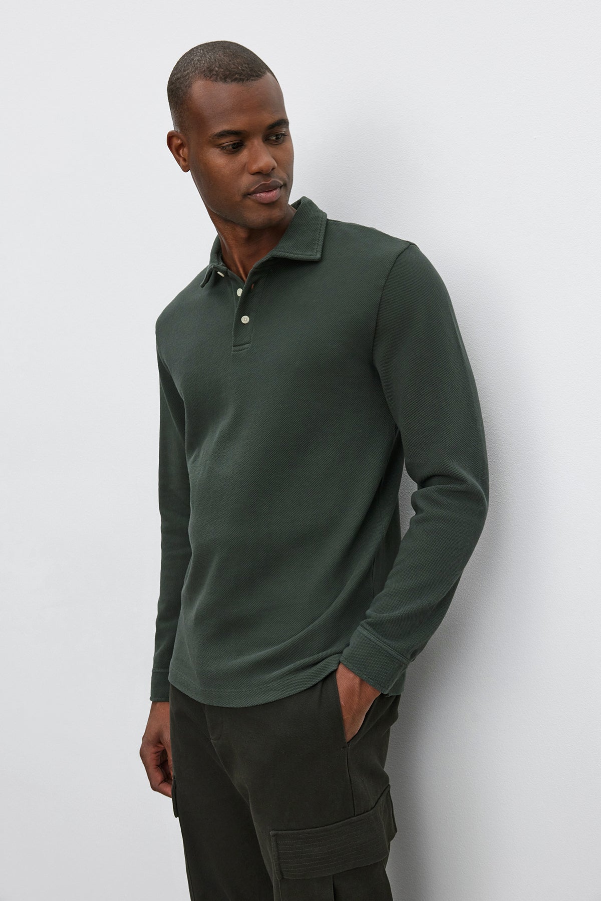   A man in a dark green MIKE POLO long-sleeve piqué cotton shirt by Velvet by Graham & Spencer and cargo pants stands against a plain white background, looking to his left with his hands in his pockets. 