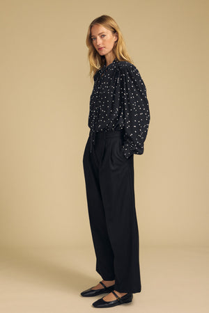 A woman stands against a beige background, wearing the ALMA SILK BLOUSE by Velvet by Graham & Spencer— a black, polka dot silk blouse—paired with black wide-leg trousers and black flat shoes.