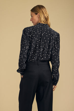 A person stands facing away, wearing the Velvet by Graham & Spencer ALMA SILK BLOUSE in black along with black trousers, set against a beige background.