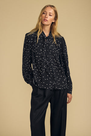 A woman stands against a beige background wearing the ALMA SILK BLOUSE by Velvet by Graham & Spencer—a black blouse with white polka dots—paired with loose black trousers, looking to the side with one hand in her pocket.