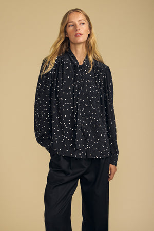 A woman wears the ALMA SILK BLOUSE by Velvet by Graham & Spencer— a loose, long-sleeve polka dot top with a bow tie neckline—paired with wide-leg black pants, standing against a beige background.