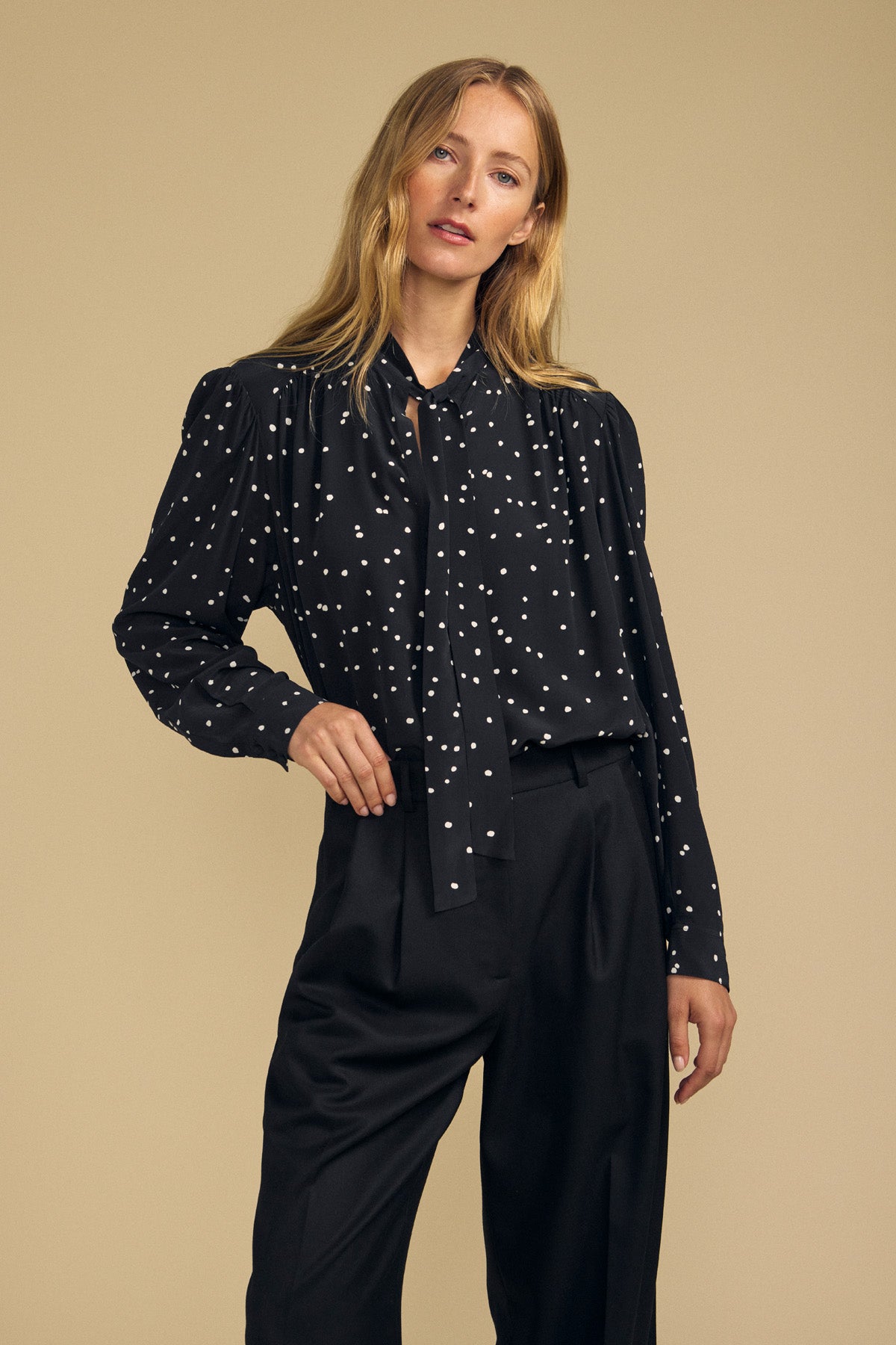 A woman stands against a beige background wearing the ALMA SILK BLOUSE by Velvet by Graham & Spencer, featuring a polka dot print, tie-neck detail, and paired with black trousers.-41935323496641
