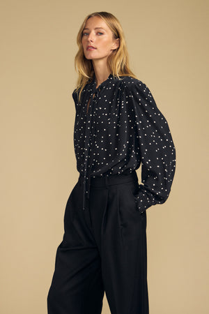 A person wearing the ALMA SILK BLOUSE by Velvet by Graham & Spencer—featuring a black fabric with white polka dots—and black high-waisted pants stands against a plain beige background.